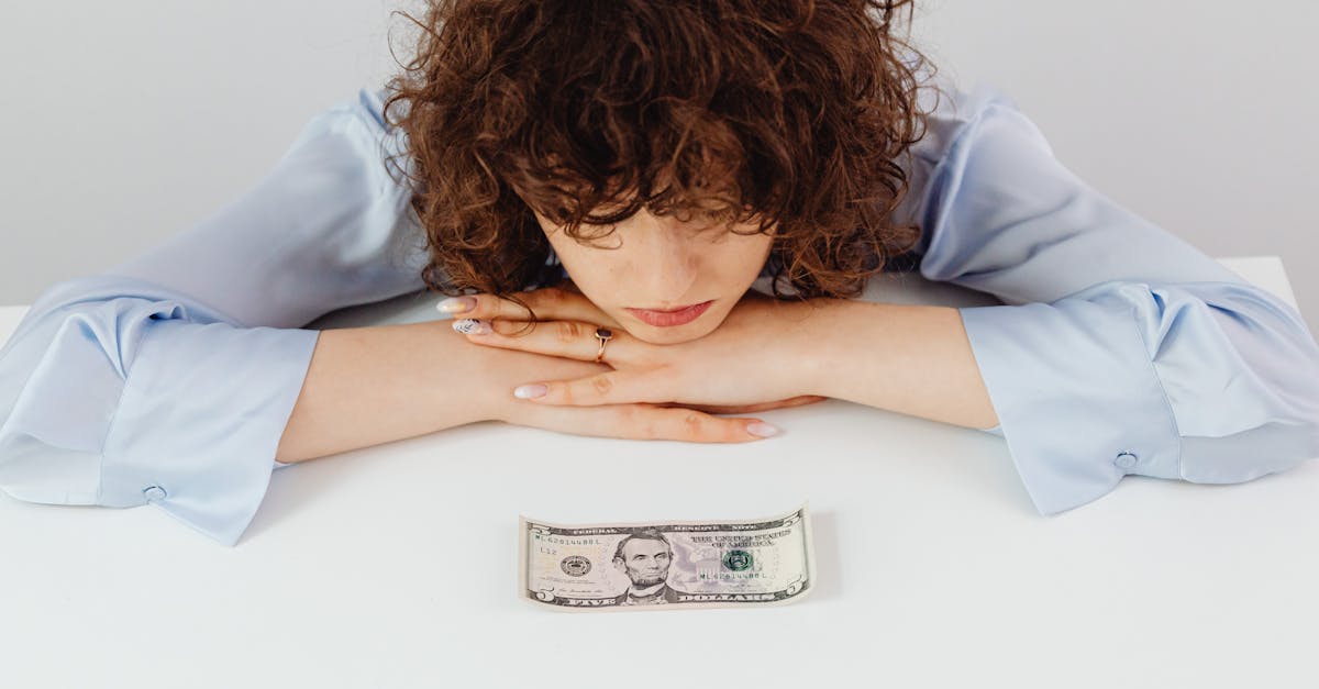 A person with curly hair looks thoughtfully at a five-dollar bill on a table