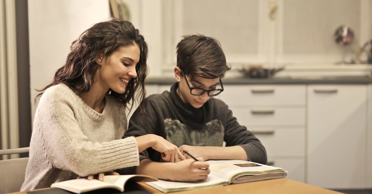 Une femme souriante aide son jeune frère ou sa jeune sœur à faire ses devoirs à la table de la cuisine, illustrant ainsi les soins et le soutien émotionnel dans un cadre familial.