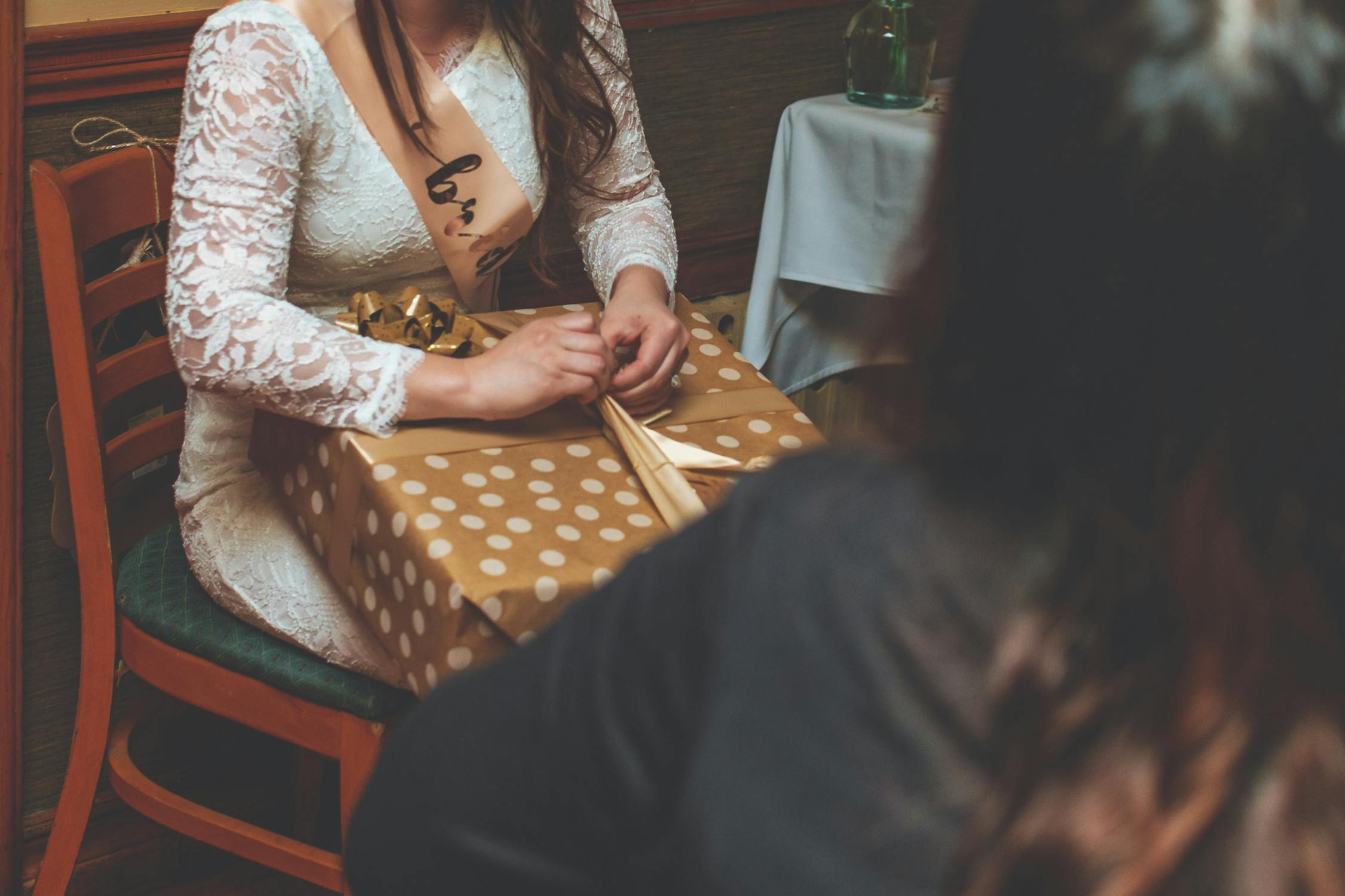 A woman in a lace dress, sitting at a table, unwraps a beautifully wrapped gift, symbolizing care and support for female cancer patients and survivors.
